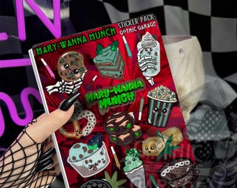 May include: A sticker pack sheet titled Mary-Wanna Munch by Gothic Garage featuring various cute, anthropomorphic food illustrations including a cookie, cake, popcorn, pretzel, ice cream, and brownies, all accented with small green cannabis leaf icons.