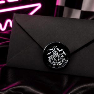 May include: A black envelope sealed with a black wax stamp featuring a Halloween design. The stamp has a pumpkin, moon, and bats in a silver colour. The background is dark with a neon pink glow.