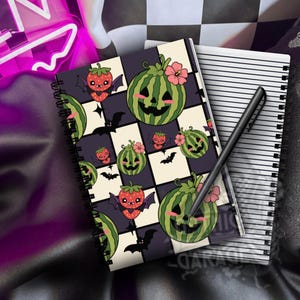 May include: A spiral-bound notebook featuring a chequered pattern of black and cream squares decorated with illustrations of smiling watermelon jack-o'-lanterns, strawberries with bat wings, and small black bats. A black pen rests on the notebook.