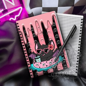 May include: A spiral-bound notebook featuring a black bat character wearing heart-shaped sunglasses and a flower, floating in a pink polka-dot pool float with skulls. The cover has pink dripping paint over black and pink stripes with skulls.