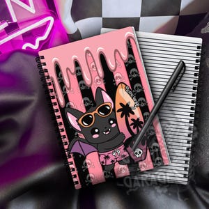 May include: A spiral-bound notebook featuring a cartoon bat wearing orange sunglasses and pink palm tree print shorts, holding a surfboard. The cover has a pink dripping effect over a black and pink striped background with skull icons and text.