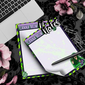 Könnte beinhalten: Zwei grün, lila und schwarz gestreifte Notizblöcke mit dem Text "Canna Mom Notes" oben. Die Notizblöcke haben ein grün, lila und schwarz gestreiftes Design mit einem grünen Marihuanablatt oben. Die Notizblöcke liegen auf einer schwarz-weißen gemusterten Oberfläche.