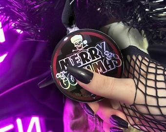Puede incluir: Adorno navideño redondo con el texto "MERRY GOTHMAS" y gráficos de calaveras. El adorno es negro con un borde rojo y una cinta negra. El fondo es de color púrpura oscuro.
