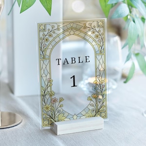 May include: Clear acrylic table number card with a floral design in yellow, green, and blue. The words "TABLE 1" are printed in black. The card is held by a small, rectangular wooden base.