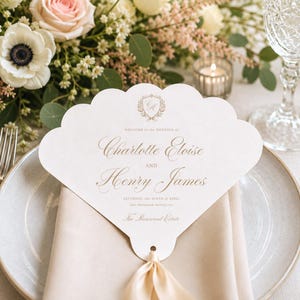 May include: A scalloped, fan-shaped wedding programme card in white with gold lettering. It features the text: Welcome to the wedding of Charlotte Eloise and Henry James, Saturday, the ninth of April, two thousand twenty-xx, The Rosewood Estate.