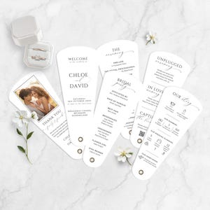 Può includere: Set di partecipazioni di nozze con ventagli di invito, una foto e anelli in una scatola. I ventagli mostrano testi come "Benvenuti", "Bridal Party" e "La nostra storia". Lo schema dei colori è bianco con testo nero, con fiori.