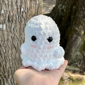 May include: A white plush ghost with black eyes and pink cheeks. The ghost is sitting in the palm of a hand.