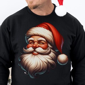 Peut inclure: Sweat-shirt noir avec une illustration détaillée du visage du Père Noël, coiffé d'un bonnet rouge et d'une longue barbe blanche. Le motif est centré sur le devant du sweat-shirt. Un sapin de Noël est en arrière-plan.