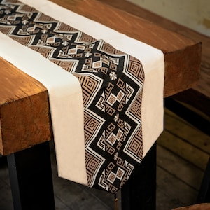 May include: A white table runner with a black and brown geometric pattern. The runner is on a wooden table with black metal legs.