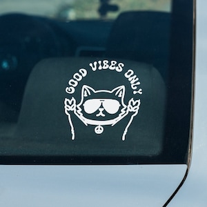 May include: A white vinyl sticker on a silver car with a cartoon cat wearing sunglasses and peace sign necklace. The sticker says "Good Vibes Only".