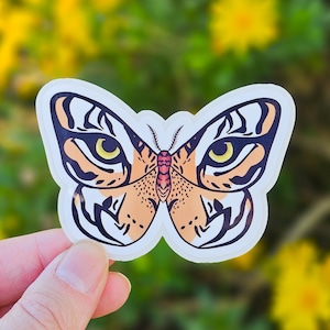 Puede incluir: Una pegatina de mariposa con un diseño único. La pegatina presenta una mariposa con un patrón similar al de un tigre en naranja, negro y blanco. Los ojos de la mariposa son amarillos. La pegatina se sostiene contra un fondo borroso de flores amarillas y follaje verde.