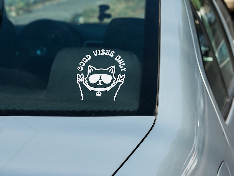 May include: A white vinyl sticker on a silver car with a cartoon cat wearing sunglasses and peace sign necklace. The sticker says "Good Vibes Only".