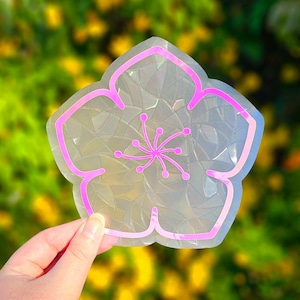 May include: A clear iridescent sticker with a pink outline of a flower. The flower has a pink center with a swirl design.