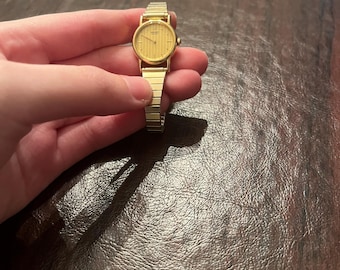 May include: A gold-toned wristwatch with a rectangular face and a matching band. The watch has a simple, elegant design with a textured dial. The band is made of small, connected links.