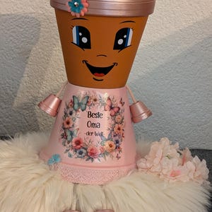Könnte beinhalten: Terrakottafarbener Blumentopf mit aufgemaltem Gesicht, rosa Kleid und zwei kleinen passenden Töpfen als Schuhe. Das Kleid hat ein Blumenmuster und den Text "Beste Oma - der Welt". Ein rosa Blumenkranz und eine flauschige weiße Oberfläche vervollständigen das Design.