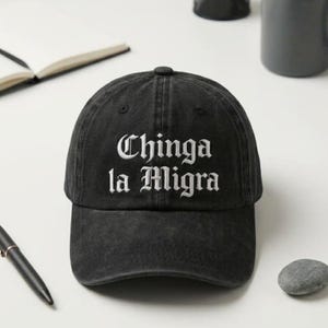 Può includere: Cappellino da baseball nero con la scritta "Chinga la Migra" ricamata in bianco. Il cappellino ha una visiera curva e un bottone superiore. Il cappello è realizzato in un materiale morbido e usurato.