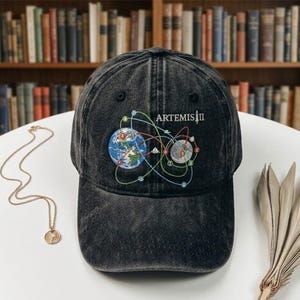 May include: A black, distressed baseball cap with an embroidered design featuring Earth, a celestial body, and the text "ARTEMIS II." The design includes colourful lines connecting the planets. The cap is sitting on a white surface.