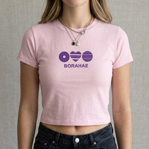 May include: A pale pink crop top featuring a purple graphic and the word "BORAHAE". The design includes a circle, a heart, and a striped shape. The top has short sleeves and a round neckline.