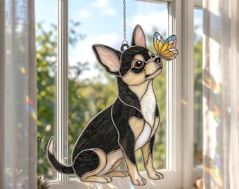 Puede incluir: Un atrapasoles de vidriera que representa a un chihuahua negro y fuego sentado con una pequeña mariposa multicolor posada en su nariz. La pieza está suspendida por una cadena de metal plateado y muestra segmentos de vidrio.