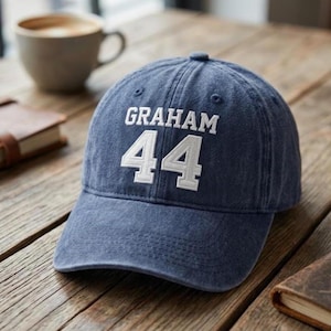 Puede incluir: Una gorra de béisbol de mezclilla azul con el texto bordado en blanco GRAHAM sobre el número 44 grande bordado en blanco. La gorra tiene una corona estructurada, una visera curva y ojales cosidos en los paneles superiores para ventilación.