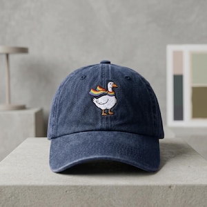 Può includere: Un cappellino da baseball blu navy con finitura invecchiata. Il cappellino presenta un disegno ricamato di un'anatra bianca con una sciarpa arcobaleno. L'anatra ha becco e zampe arancioni. Il cappellino è su una superficie grigia.