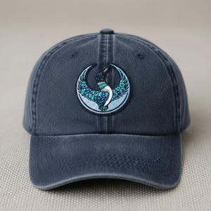 以下が含まれることがあります： ネイビーブルーの野球帽。鳥のデザインが刺繍されたパッチが付いています。パッチは円形で、三日月と青、緑、白のアクセントが施された鳥のデザインです。帽子はカーブしたつばと布製のストラップが付いています。