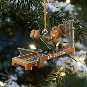 Könnte beinhalten: Ein hängender Weihnachtsbaumschmuck in Form einer braunen Lebkuchenfigur mit Pferdeschwanz, die in einem grünen Sport-BH und Leggings auf einem Pilates-Reformer aus Holz trainiert. An der Seite des Holzrahmens steht der Text EMILY 2026 gedruckt.