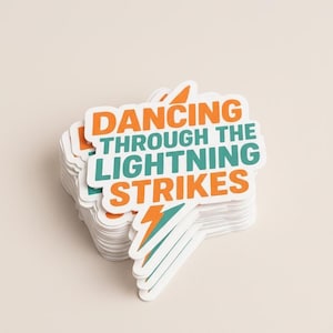 May include: A stack of white stickers with the text "DANCING THROUGH THE LIGHTNING STRIKES" in orange and teal. The design includes a lightning bolt graphic. The stickers have a white border and are on a light beige background.