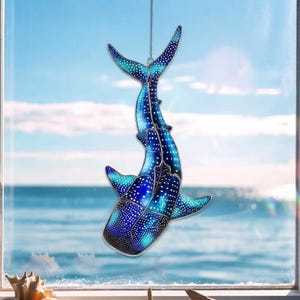 Puede incluir: Un atrapasueños de cristal tintado con forma de tiburón ballena, que presenta un degradado azul vibrante con patrones de puntos blancos. La pieza está construida con varios segmentos de vidrio unidos por líneas metálicas oscuras y cuelga de una cadena.