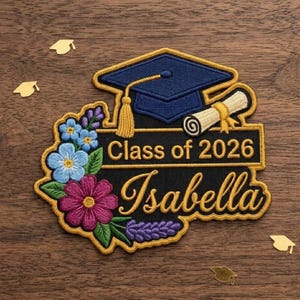Pode incluir: Patch bordado com tema de formatura. Apresenta um chapéu de formatura azul marinho, um pergaminho e o texto "Class of 2026 Isabella". O patch é cercado por bordados florais coloridos em tons de azul, rosa e roxo, com uma borda dourada.