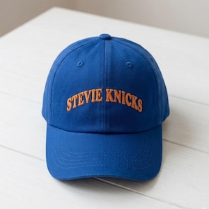 May include: A royal blue baseball cap featuring the text STEVIE NICKS embroidered in orange thread across the front panel. The cap has a structured crown, a curved brim, and a button at the top center, showcasing a classic six-panel design.