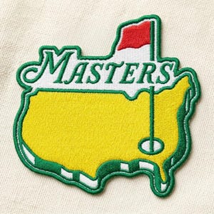 Puede incluir: Parche bordado con la palabra "MASTERS" en escritura verde sobre un contorno de mapa amarillo y verde con una bandera roja. El parche tiene un borde verde y blanco, y es probablemente para los entusiastas del golf.