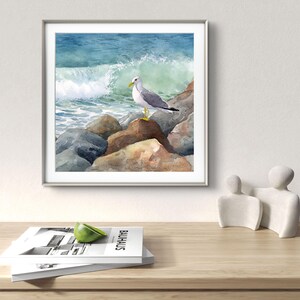 May include: Watercolor painting of a seagull perched on rocks at the edge of the ocean. The artwork features a crashing wave and a blue and green color palette, framed in a silver frame. The painting is displayed on a wooden table.