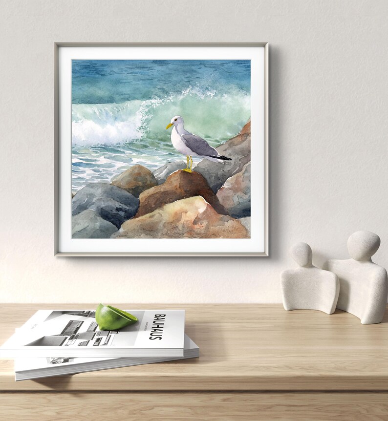 May include: Watercolor painting of a seagull perched on rocks at the edge of the ocean. The artwork features a crashing wave and a blue and green color palette, framed in a silver frame. The painting is displayed on a wooden table.
