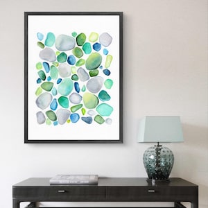 Puede incluir: Una impresi&oacute;n en acuarela enmarcada con un dise&ntilde;o abstracto de formas redondeadas y coloridas en tonos verdes, azules y grises. La obra de arte se exhibe sobre un fondo blanco dentro de un marco negro. La impresi&oacute;n est&aacute; sobre una mesa negra con una l&aacute;mpara.