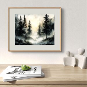 May include: A framed watercolor painting of a misty forest scene. Tall, dark evergreen trees are silhouetted against a soft, golden light. The painting is in a light wood frame, resting on a wooden surface with a stack of books and a decorative sculpture.