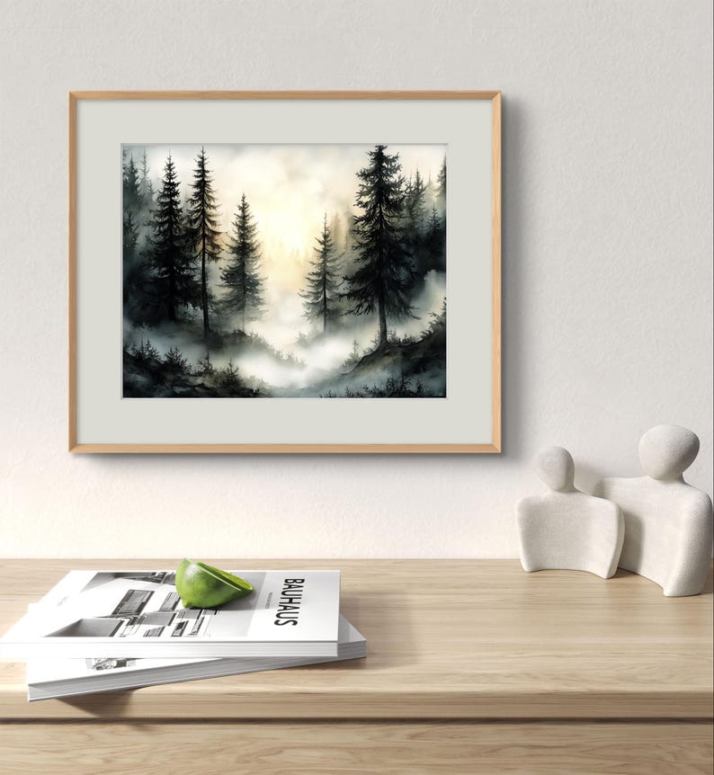 May include: A framed watercolor painting of a misty forest scene. Tall, dark evergreen trees are silhouetted against a soft, golden light. The painting is in a light wood frame, resting on a wooden surface with a stack of books and a decorative sculpture.