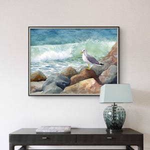May include: Watercolor painting of a seagull perched on rocks at the ocean's edge. The artwork depicts a crashing wave and a blue-green sea. The framed art piece is displayed above a dark table with a lamp.