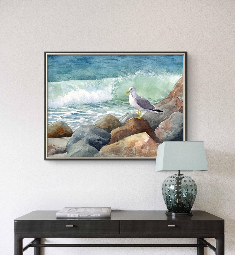 May include: Watercolor painting of a seagull perched on rocks at the ocean's edge. The artwork depicts a crashing wave and a blue-green sea. The framed art piece is displayed above a dark table with a lamp.