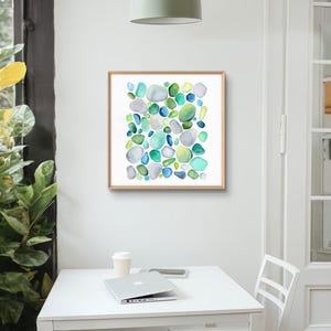 Puede incluir: Una impresi&oacute;n en acuarela enmarcada con un dise&ntilde;o abstracto de formas redondeadas y coloridas en tonos azules, verdes y grises. La obra de arte se muestra en un marco de madera clara. La impresi&oacute;n se muestra en una habitaci&oacute;n con una mesa blanca y un port&aacute;til.
