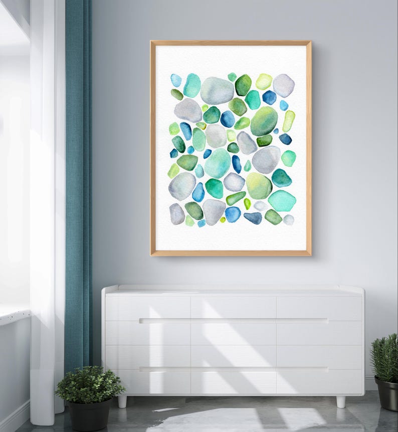 Puede incluir: Una impresi&oacute;n en acuarela enmarcada que presenta un dise&ntilde;o abstracto de piedras redondeadas y coloridas. Las piedras son en tonos de azul, verde y gris, sobre un fondo blanco. El marco es de madera clara. La obra de arte se exhibe sobre una c&oacute;moda blanca.
