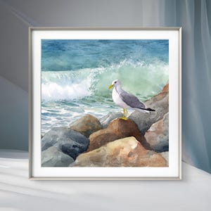 May include: Watercolor painting of a seagull perched on rocks at the ocean's edge. The artwork features a white and gray seagull with a yellow beak and legs, set against a backdrop of crashing waves and a blue-green sea. The painting is framed.