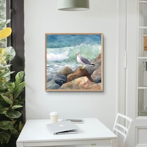 May include: Watercolor painting of a seagull perched on rocks at the edge of the ocean. The artwork features a wooden frame and depicts a crashing wave with shades of blue and green. The seagull is in the center of the image.