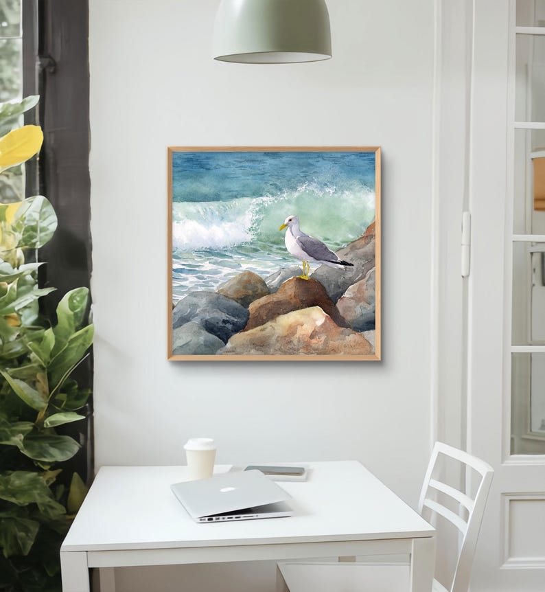 May include: Watercolor painting of a seagull perched on rocks at the edge of the ocean. The artwork features a wooden frame and depicts a crashing wave with shades of blue and green. The seagull is in the center of the image.