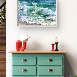 May include: A light green chest of drawers with six drawers and gold-coloured handles, topped with a red curved lamp, two candle holders, and a framed painting of ocean waves crashing onto a sandy shore with shades of blue, turquoise, and white foam.