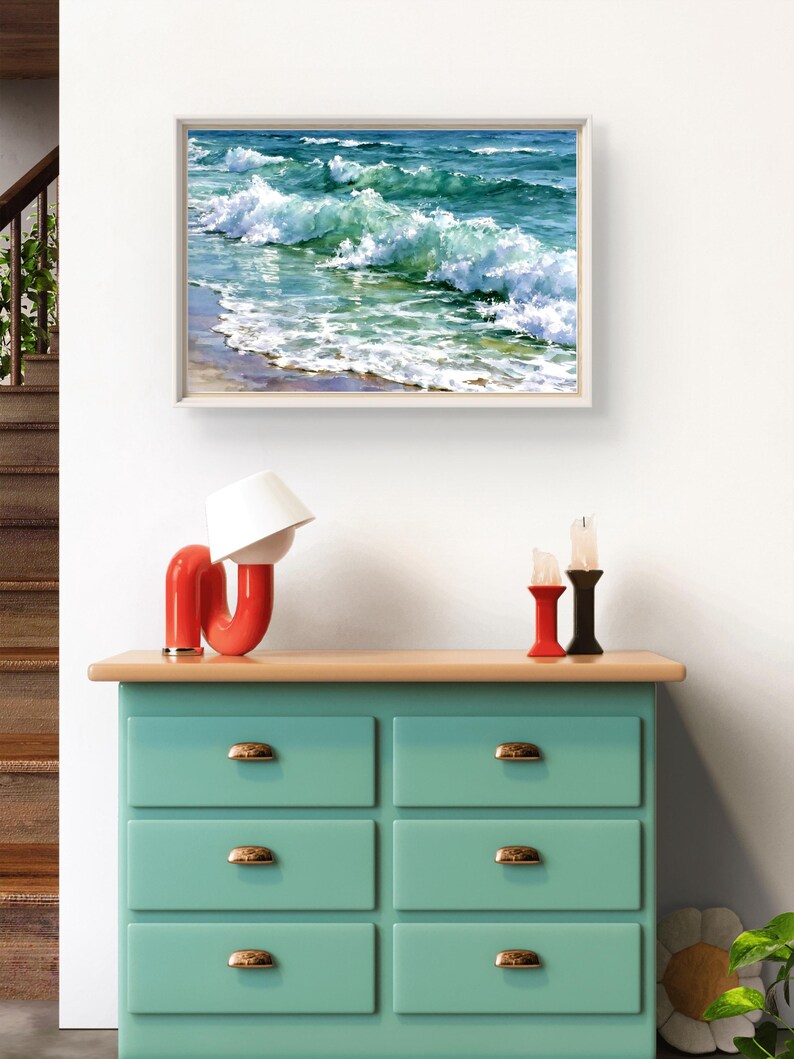 May include: A light green chest of drawers with six drawers and gold-coloured handles, topped with a red curved lamp, two candle holders, and a framed painting of ocean waves crashing onto a sandy shore with shades of blue, turquoise, and white foam.