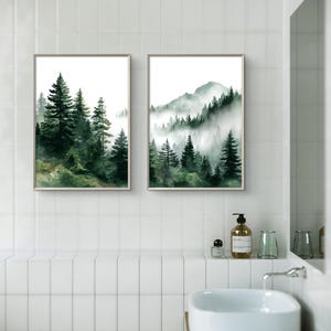 May include: Two framed watercolor prints of forest scenes. The left print features tall evergreen trees, while the right print depicts a mountain range with trees shrouded in mist. The prints have a neutral frame and are displayed in a bathroom setting.