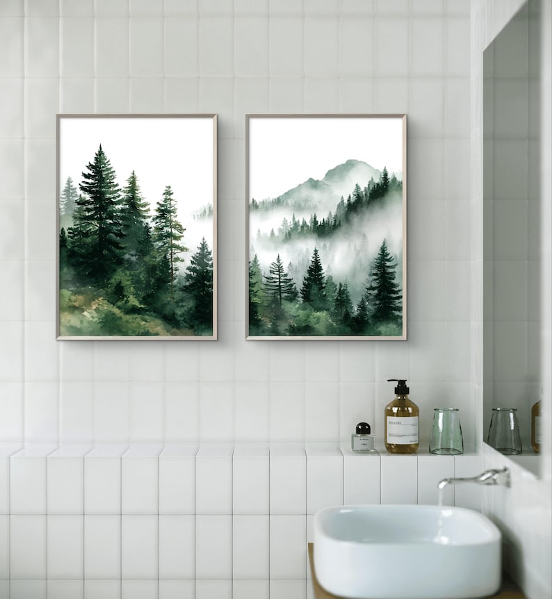 May include: Two framed watercolor prints of forest scenes. The left print features tall evergreen trees, while the right print depicts a mountain range with trees shrouded in mist. The prints have a neutral frame and are displayed in a bathroom setting.