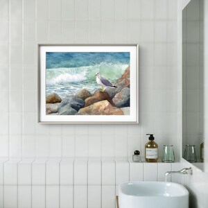 May include: A framed watercolor painting of a seagull perched on rocks near a crashing ocean wave. The artwork features shades of blue, green, and brown, with a silver frame. The painting is displayed in a bathroom setting.