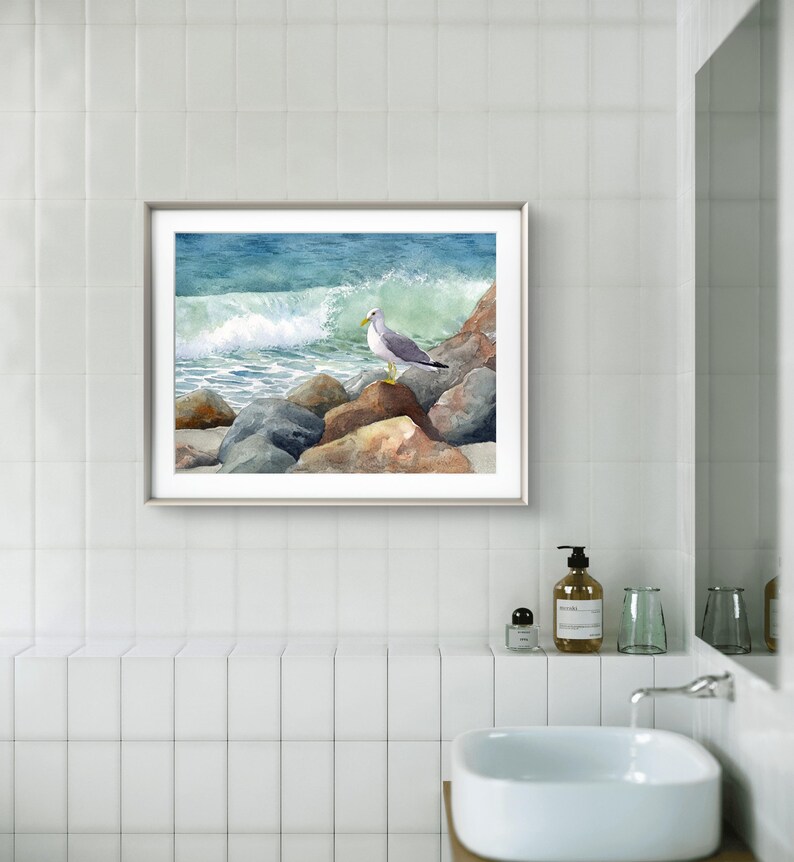 May include: A framed watercolor painting of a seagull perched on rocks near a crashing ocean wave. The artwork features shades of blue, green, and brown, with a silver frame. The painting is displayed in a bathroom setting.